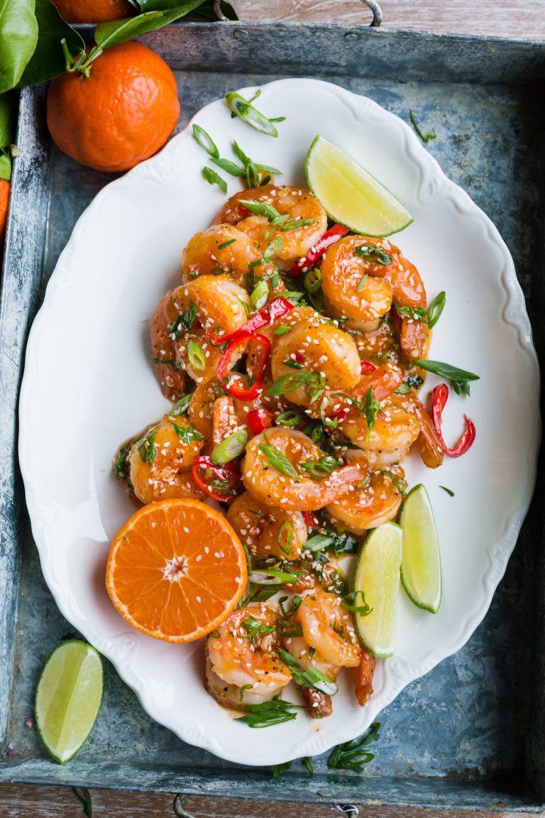 An overhead image of sweet chili sesame shrimp served on a white platter, garnished with lime wedges, peppers, and scallions.