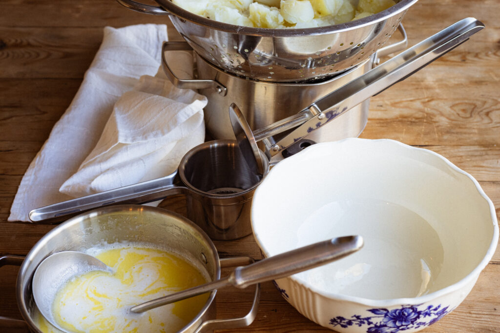 Ingredients and supplies needed to make fluffy mashed potatoes -- soft boiled potatoes, a potato masher, a mixture of cream and butter, and a large serving dish.