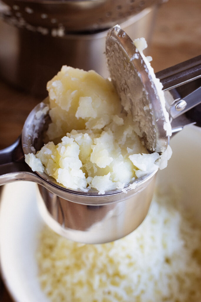 Chunks of tender boiled potatoes packed into a potato ricer to make smooth and creamy mashed potatoes.