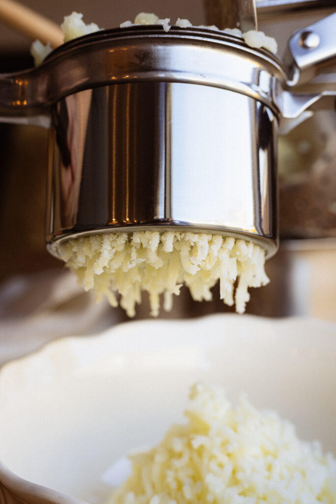 Boiled potatoes pressed through a potato ricer fall into a bowl.