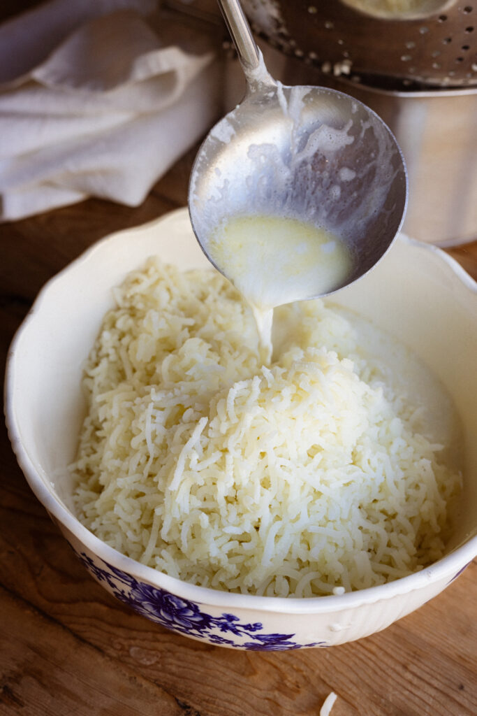 A person pours a ladle of melted butter and cream over the riced potatoes to make a creamy, buttery mashed potato mixture.