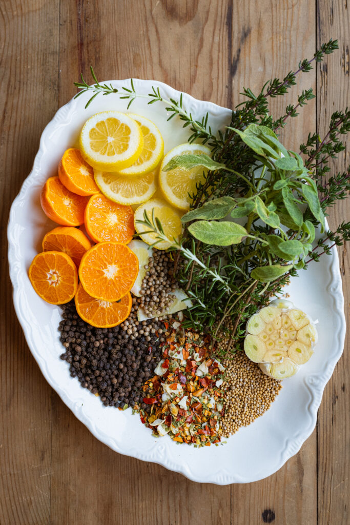 An overhead image of a plate of ingredients for an easy organic turkey brine recipe. The ingredients include fresh lemon and orange slices, sprigs of fresh herbs, peppercorns, spices, and a head of garlic.