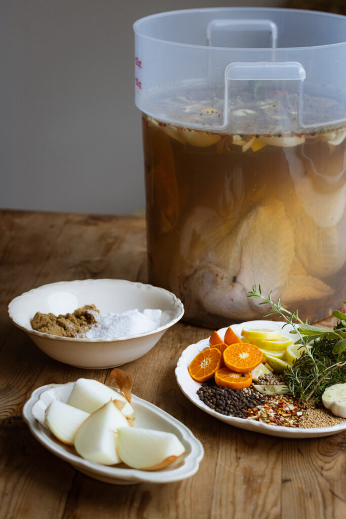 Three small plates of ingredients surround a large, clear Cambro bucket holding a raw turkey soaking in a brining solution.